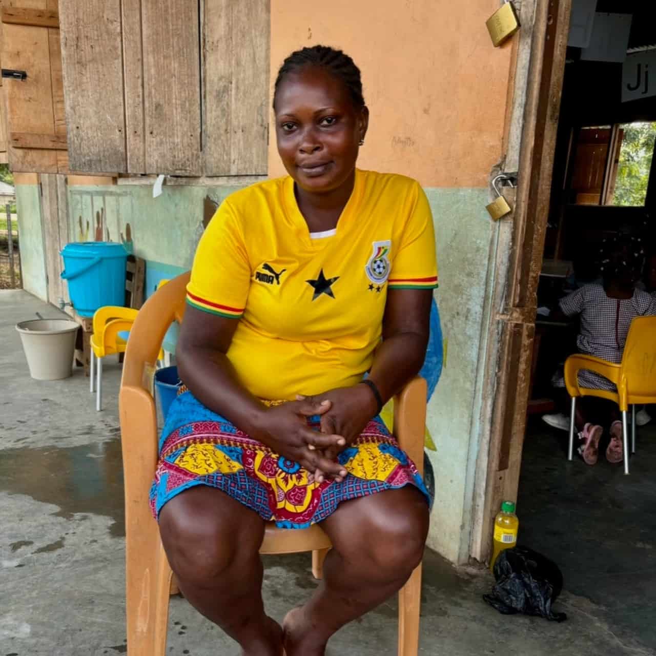 Mercy in a yellow tshirt and colourful skirt sitting in a chair in front of the creche
