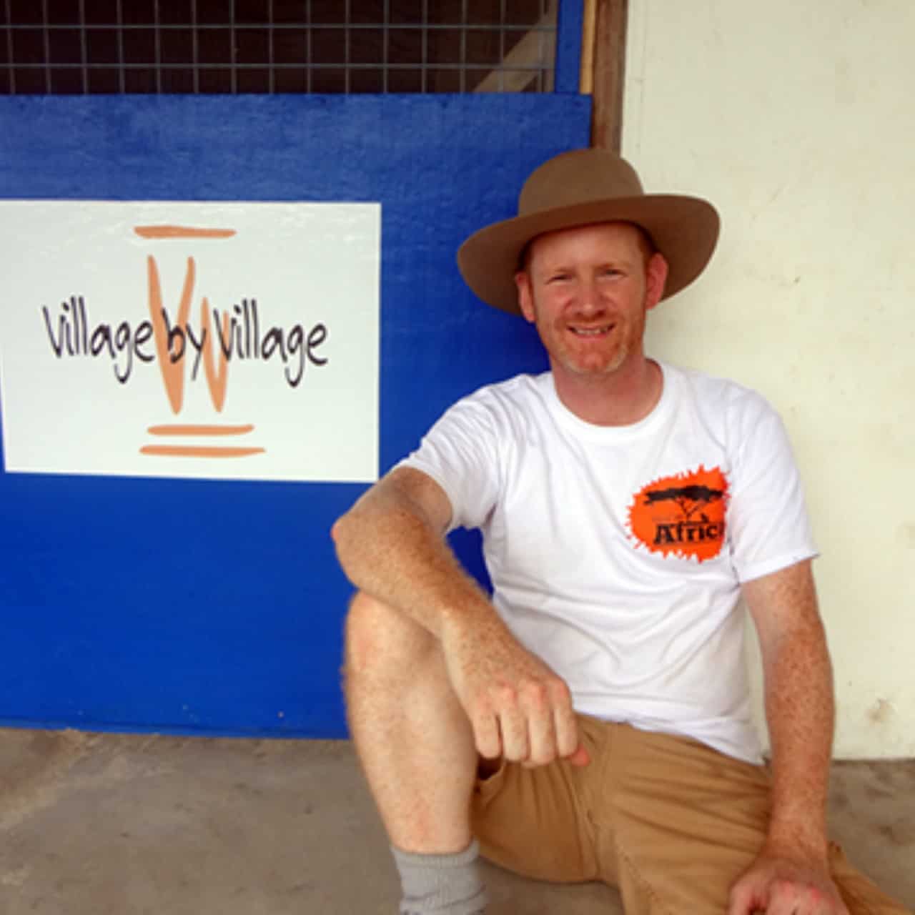 Dave sitting by the old Village by Village logo in shorts, a white tee, and a wide brimmed hat